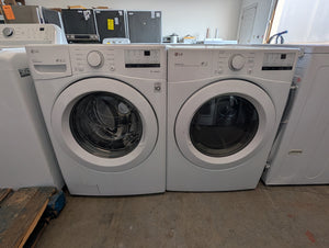 A pair of white LG front-load washing machines side by side in a showroom or warehouse setting with other appliances visible in the background.